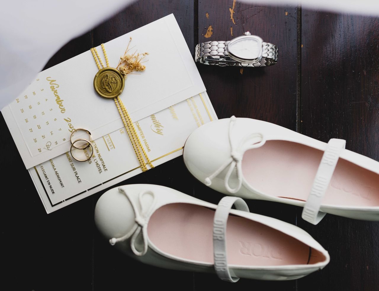 A stylish flat lay of wedding essentials including shoes, invitation, rings, and a watch on dark wood.